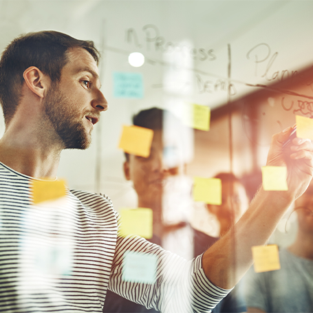 Blick durch eine Glasscheibe auf einen Mann, der auf ein Post-it auf der Scheibe schreibt. Eine Gruppe Menschen schaut ihm zu.